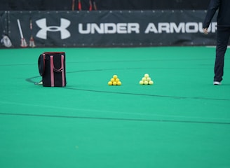 A sports field features a black and pink sports bag, two pyramids of yellow balls, and a portion of a person in dark athletic gear. The background displays a banner with the Under Armour logo.