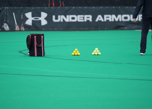 A sports field features a black and pink sports bag, two pyramids of yellow balls, and a portion of a person in dark athletic gear. The background displays a banner with the Under Armour logo.