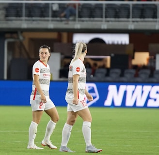 Soccer players in team uniforms stand on a grass field with a large digital advertisement screen displaying the acronym 'NWSL' in the background. The setting appears to be a stadium with rows of empty seats visible above the screen.