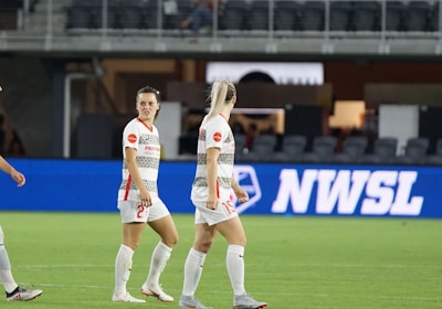 Soccer players in team uniforms stand on a grass field with a large digital advertisement screen displaying the acronym 'NWSL' in the background. The setting appears to be a stadium with rows of empty seats visible above the screen.