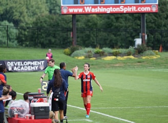 A soccer match scene with a player in a red and blue uniform walking off the field, high-fiving a staff member near the sidelines. A substitution board is held by an official dressed in green. In the background, a scoreboard displays the score, and there is a sign for the Washington Spirit, with greenery and a fence visible further back.