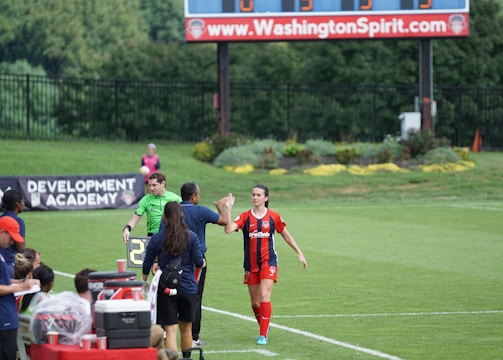 A soccer match scene with a player in a red and blue uniform walking off the field, high-fiving a staff member near the sidelines. A substitution board is held by an official dressed in green. In the background, a scoreboard displays the score, and there is a sign for the Washington Spirit, with greenery and a fence visible further back.