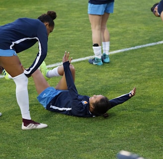 A group of athletes in sportswear are on a green soccer field. Two individuals are interacting closely; one helps the other who is lying on the grass. Nearby, other athletes are stretching.