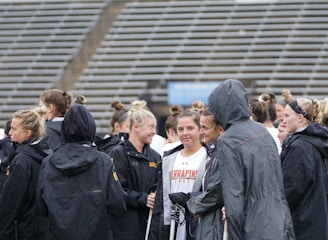 A group of people, likely athletes, are gathered together on a sports field. They are wearing black jackets with some individuals dressed in white sports uniforms underneath. A few people have their hair tied up in buns, and most are engaged in conversation. The background shows tiered stadium seats, suggesting the setting is an outdoor sports venue.