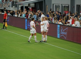 Two soccer players in white uniforms stand on the sideline of a field near a digital advertising board. One player holds a beverage bottle, and they appear to be in conversation. A referee in black stands nearby, and spectators fill the stands in the background, watching the event.