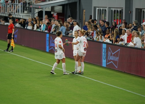 Two soccer players in white uniforms stand on the sideline of a field near a digital advertising board. One player holds a beverage bottle, and they appear to be in conversation. A referee in black stands nearby, and spectators fill the stands in the background, watching the event.