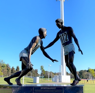 A sculpture depicting two athletes in mid-action is prominently displayed against a clear blue sky. One figure is reaching out to help the other, illustrating a moment of sportsmanship. The bronze statues stand on a black pedestal with an inscription, situated in an open green field with trees and sports facilities in the background. A tall floodlight tower looms overhead, adding to the outdoor sports setting.