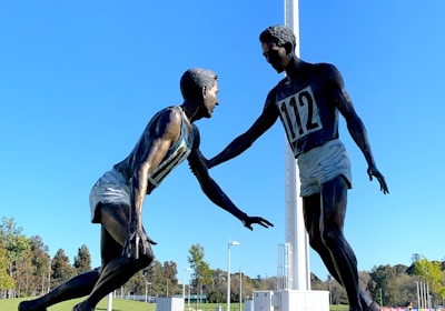 A sculpture depicting two athletes in mid-action is prominently displayed against a clear blue sky. One figure is reaching out to help the other, illustrating a moment of sportsmanship. The bronze statues stand on a black pedestal with an inscription, situated in an open green field with trees and sports facilities in the background. A tall floodlight tower looms overhead, adding to the outdoor sports setting.
