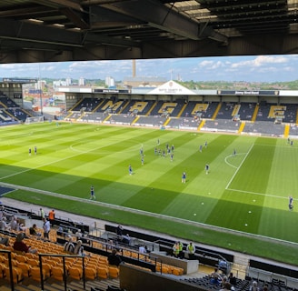 A sports stadium with a well-maintained grass field is depicted. Players are spread across the field, engaging in warm-up activities. The stands are partially filled with spectators, and the stadium is surrounded by urban buildings under a partly cloudy sky.