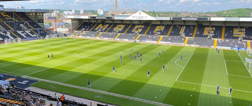 A sports stadium with a well-maintained grass field is depicted. Players are spread across the field, engaging in warm-up activities. The stands are partially filled with spectators, and the stadium is surrounded by urban buildings under a partly cloudy sky.