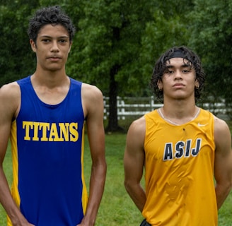 Two young athletes wearing sleeveless sports tops stand on a grassy field. The athlete on the left is wearing a blue top with the word 'TITANS' while the one on the right wears a yellow top with 'ASIJ'. The background features green trees and a white fence, with a bench visible on the right.
