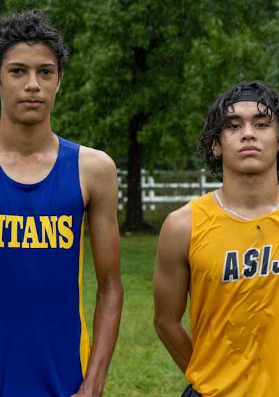 Two young athletes wearing sleeveless sports tops stand on a grassy field. The athlete on the left is wearing a blue top with the word 'TITANS' while the one on the right wears a yellow top with 'ASIJ'. The background features green trees and a white fence, with a bench visible on the right.